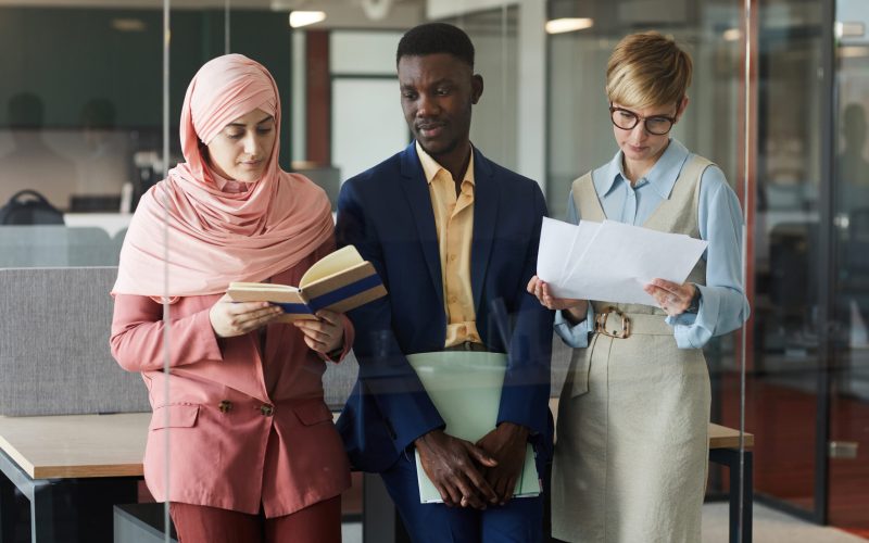 Waist up portrait of modern multi-ethnic business team of three holding documents while standing behind glass wall in office